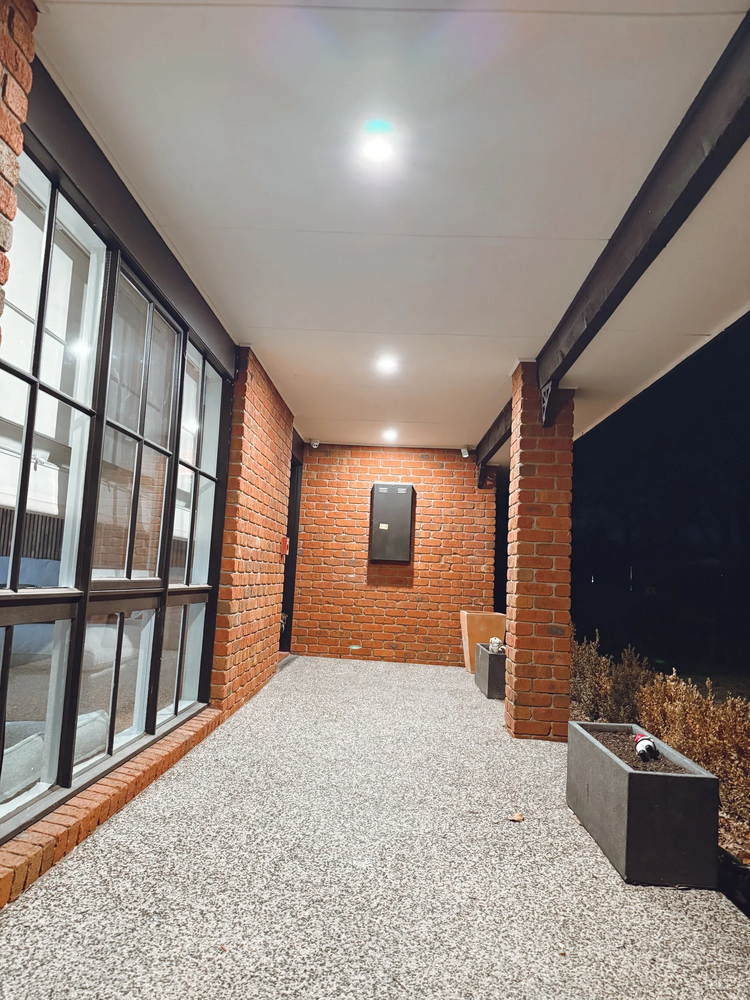 Night view of a covered outdoor patio with brick walls, granite flooring, large windows, and ceiling lights.