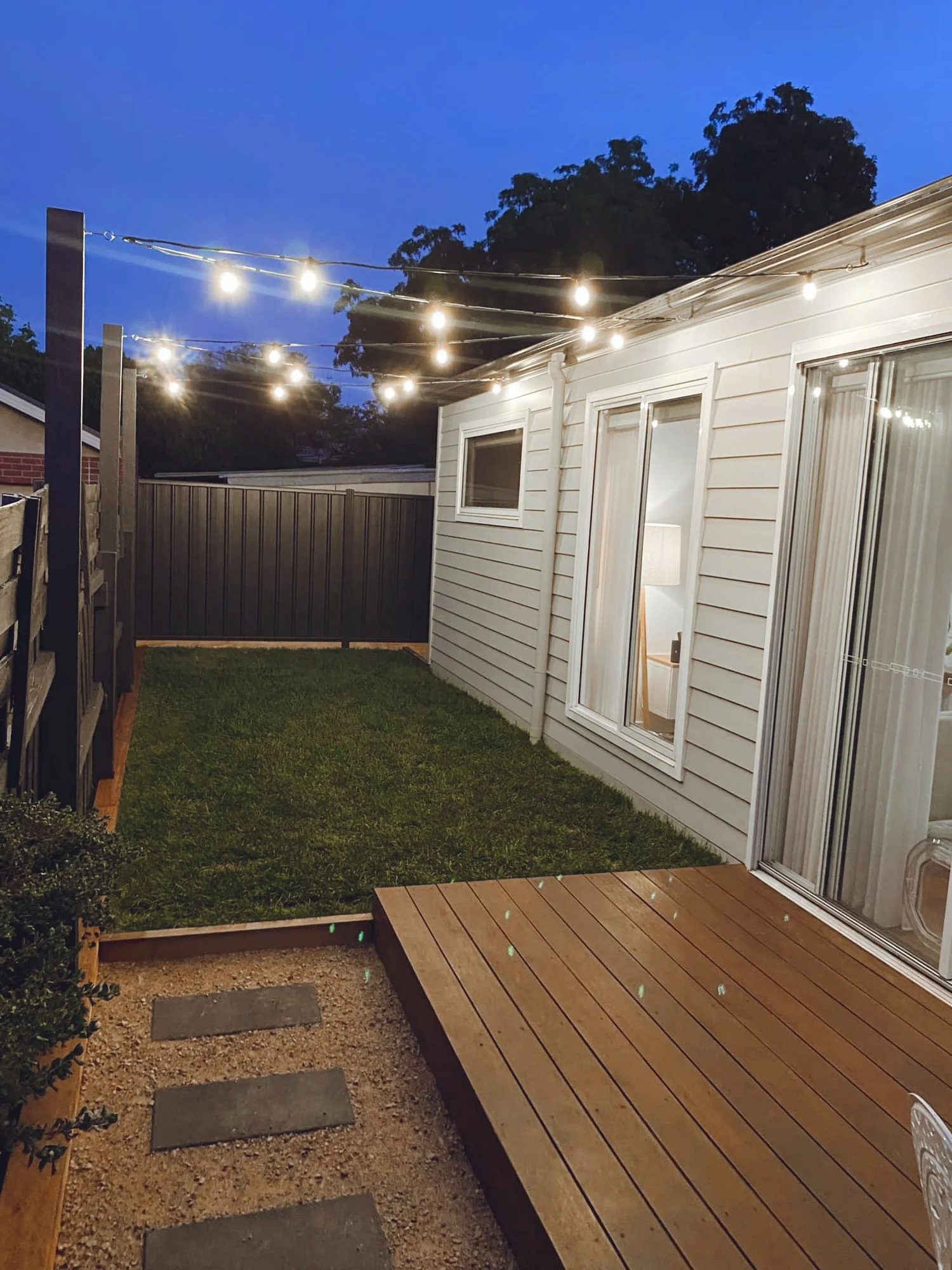 A backyard patio at dusk with string lights overhead, a sliding glass door, and a small wooden deck next to a grassy area surrounded by a wooden boundary and fence.