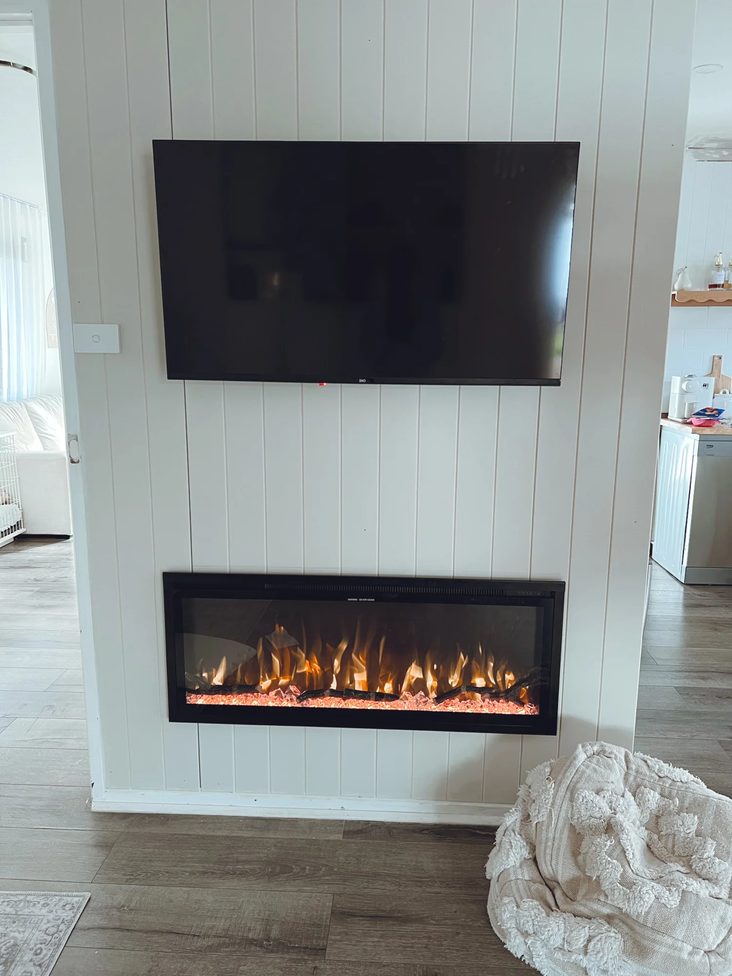 Wall-mounted flat-screen TV above an electric fireplace with realistic flames, on a white paneled wall in a living room. A beige textured throw blanket is on the floor in front of the fireplace.