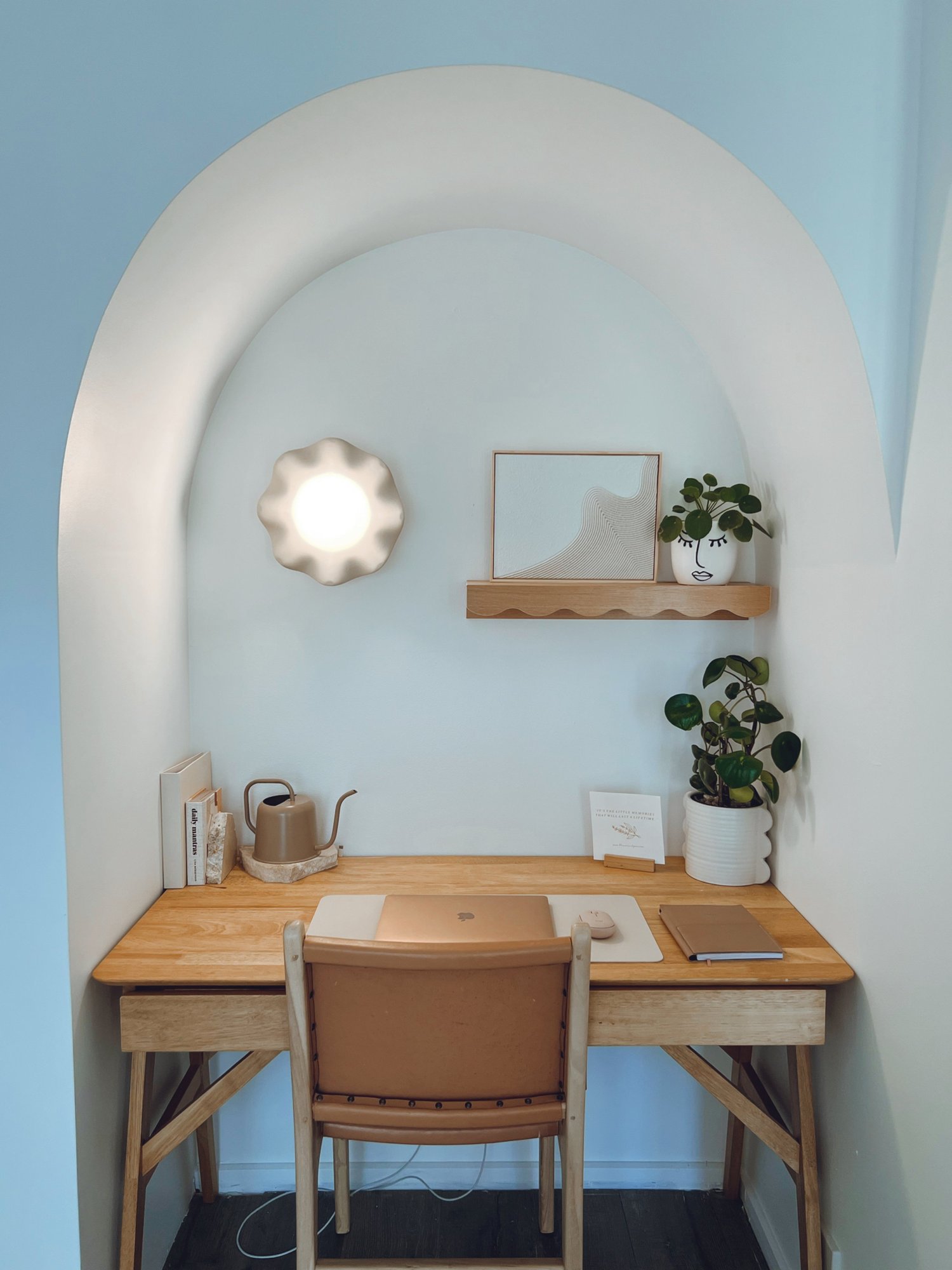 A minimalist home office setup in a small alcove with an arched ceiling. It features a wooden desk, a beige chair, a laptop, a mouse, and a closed notebook. On the desk, there are a few books, a beige watering can, and some decor. A small wooden shel