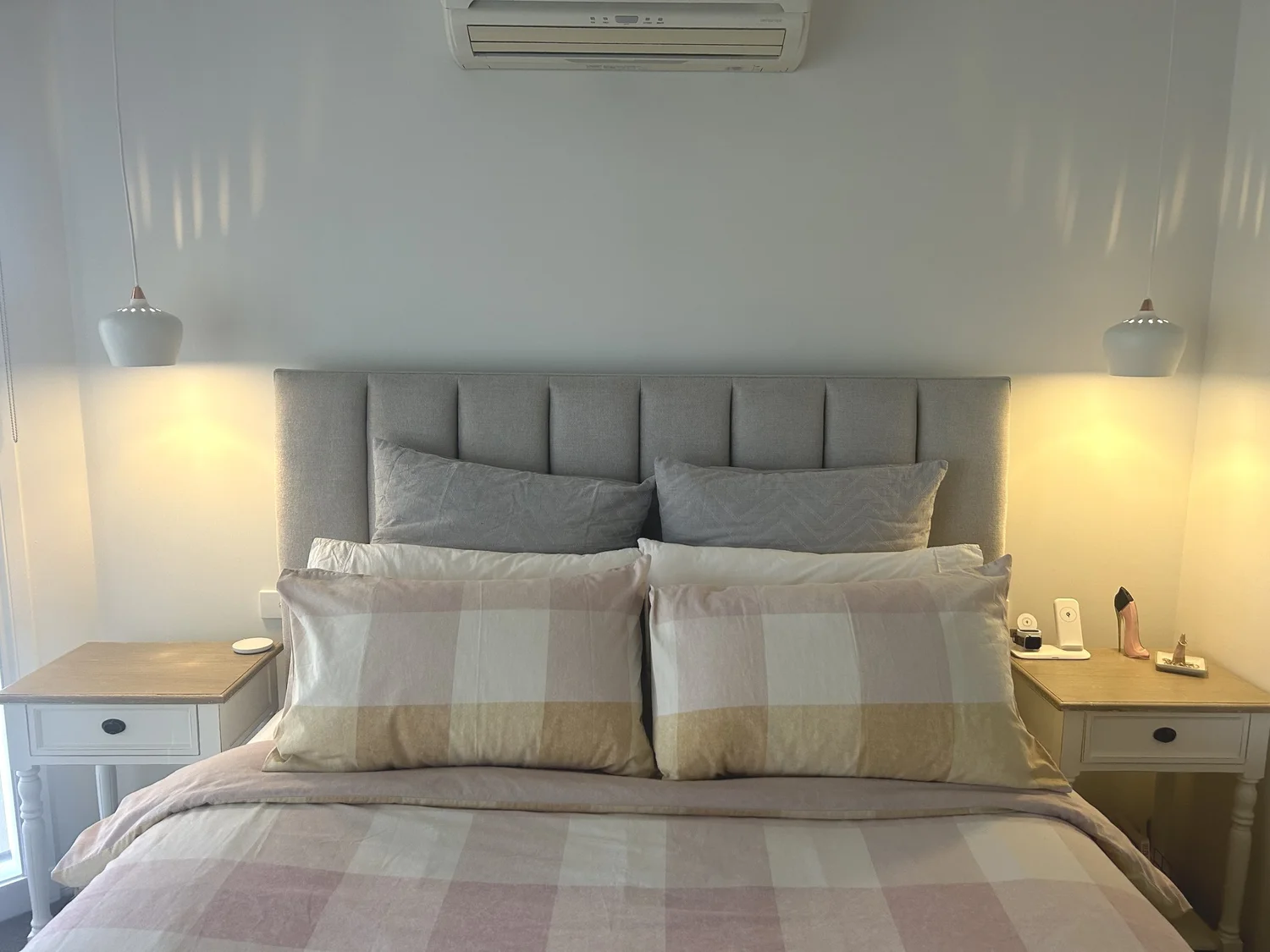 A neatly made bed with a padded gray headboard, featuring large gray and cream checked pillows and a beige, pink, and cream bedspread. Two white nightstands with drawers are on each side of the bed, with hanging pendant lamps above. On the right nigh