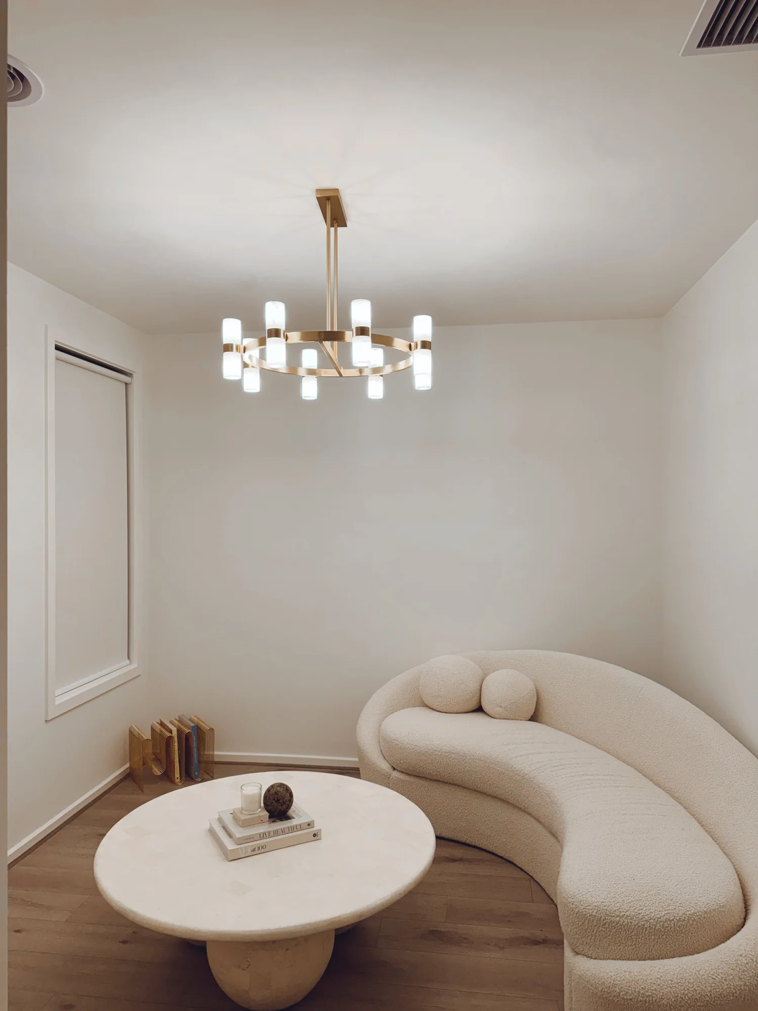 Minimalist living room with curved cream sofa, round marble coffee table, modern chandelier, and wooden floor.
