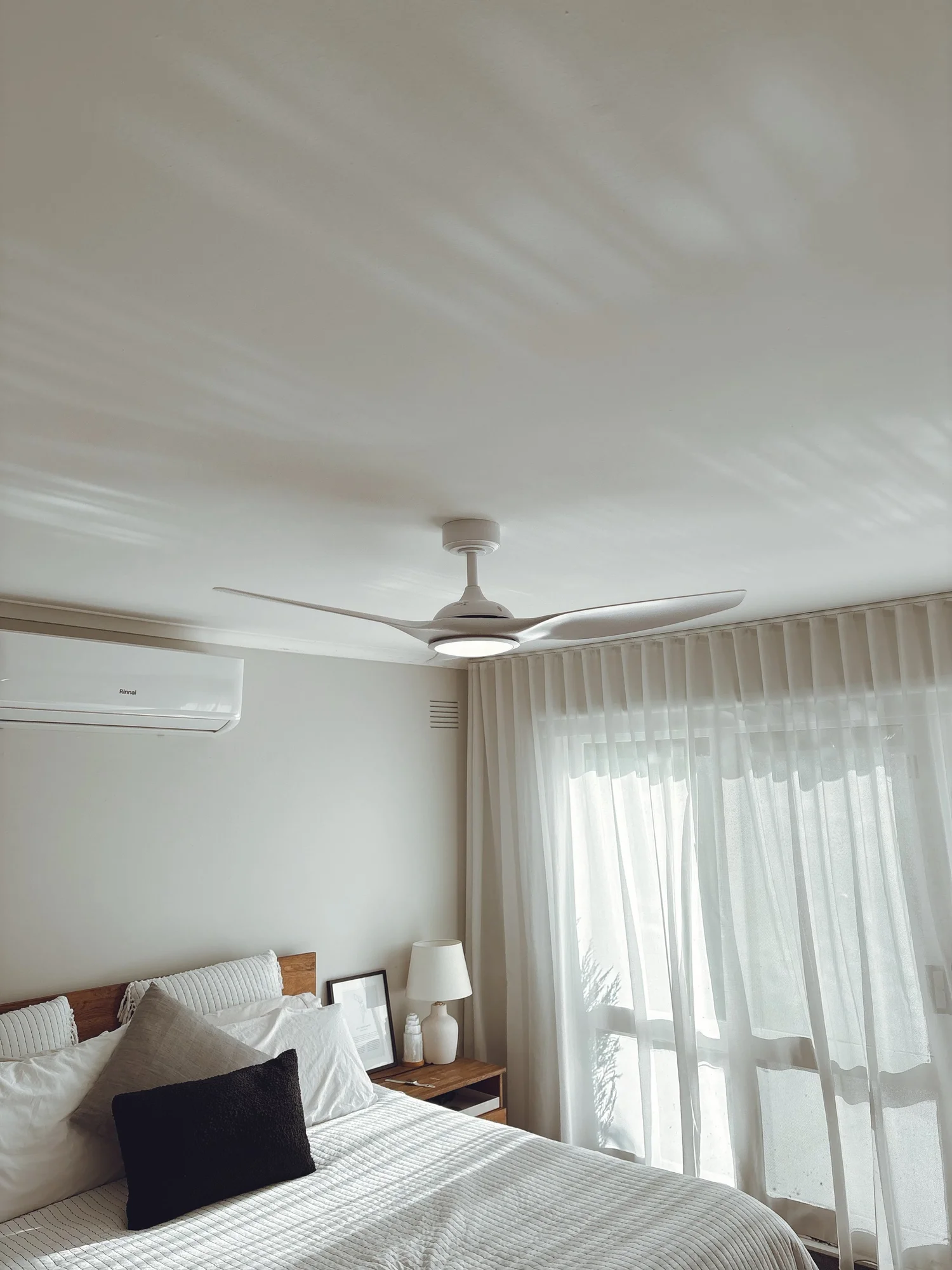 A bedroom with a bed, bedside table, lamp, framed picture, and sheer curtains in front of a large window.