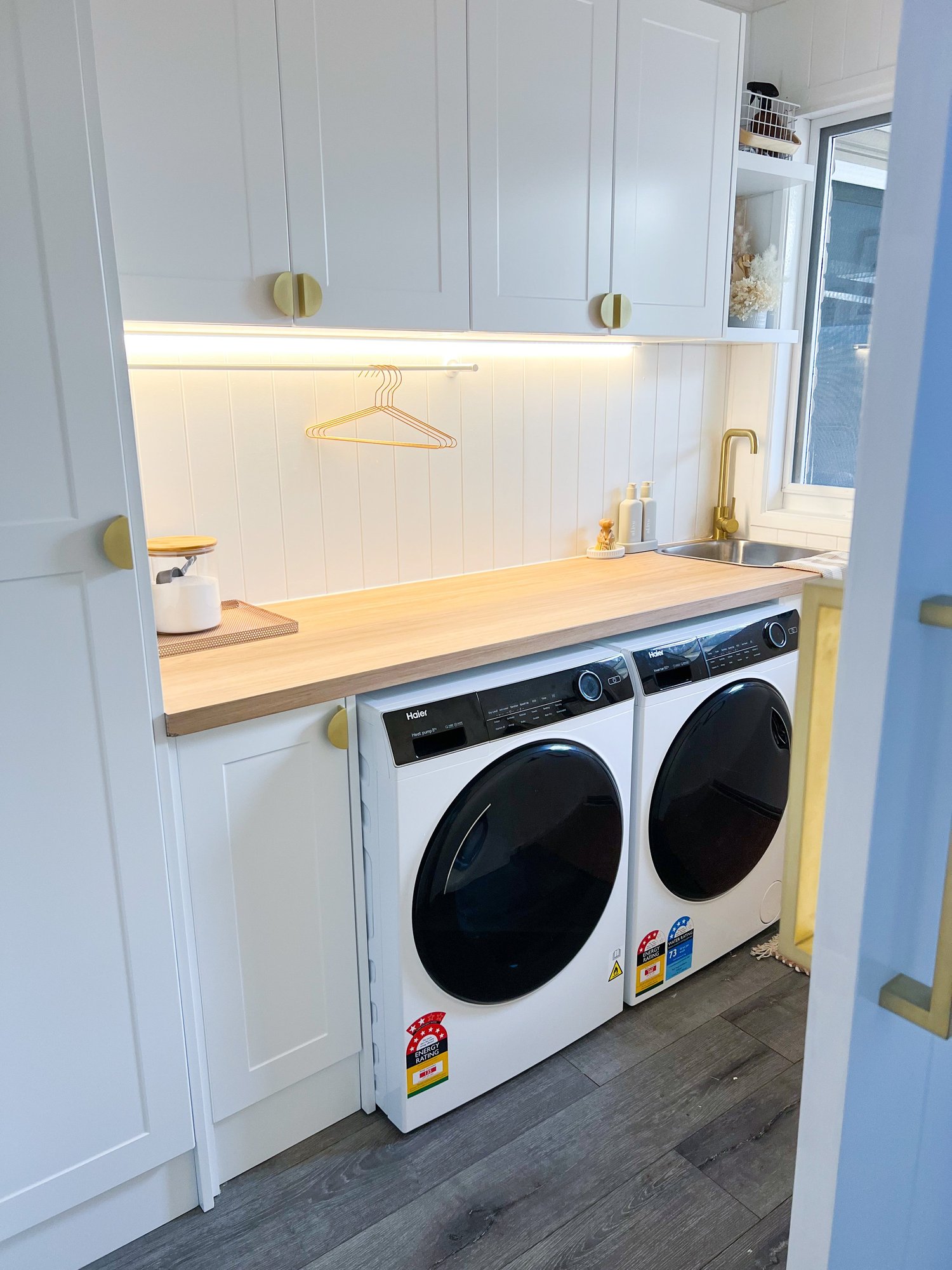 A laundry room with a wooden countertop, white cabinets with yellow knobs, a window, and a washer and dryer, all in a modern style with minimal decor.