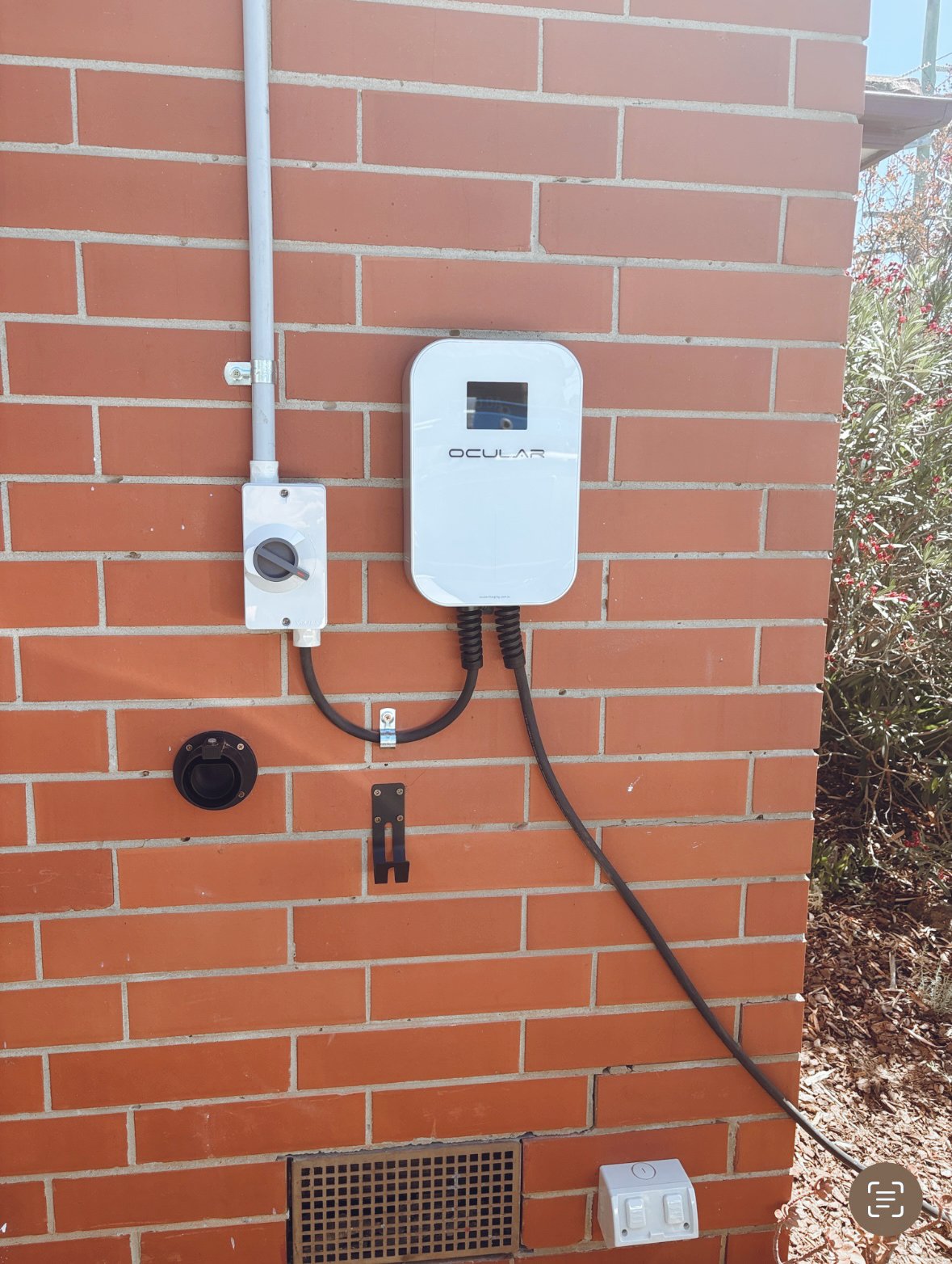 Outdoor wall-mounted solar power system with a white solar inverter labeled 'Ocular', a black cable, an electrical box, a ground vent, and an outdoor power outlet on a red brick wall.