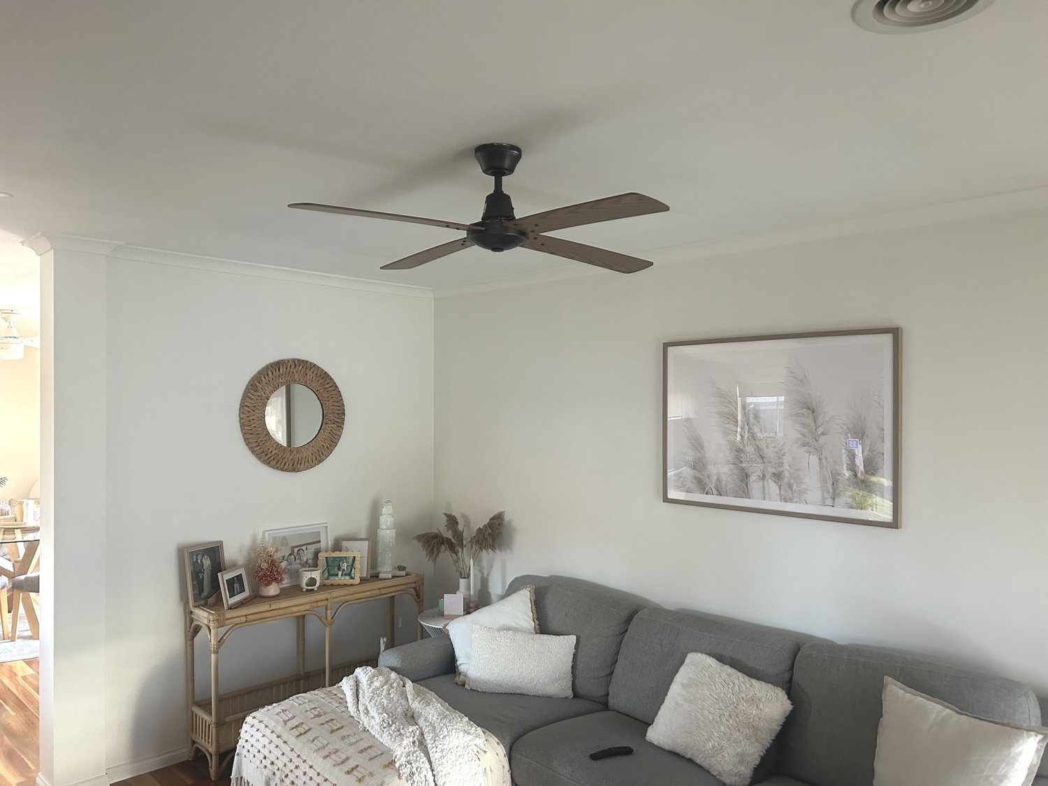 Living room with a gray sectional sofa, white pillows, wall art of pampas grass, a wicker mirror, and a decorative console table with framed photos, a vase, and dried plants.