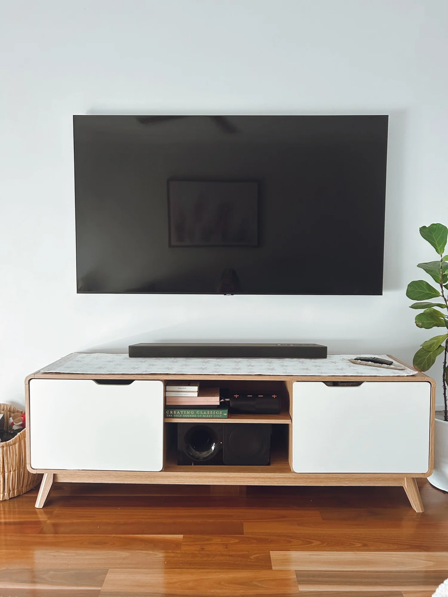 A modern wooden TV stand with white cabinet doors and open shelving, holding books and audio equipment, placed on a polished wood floor next to a potted plant.